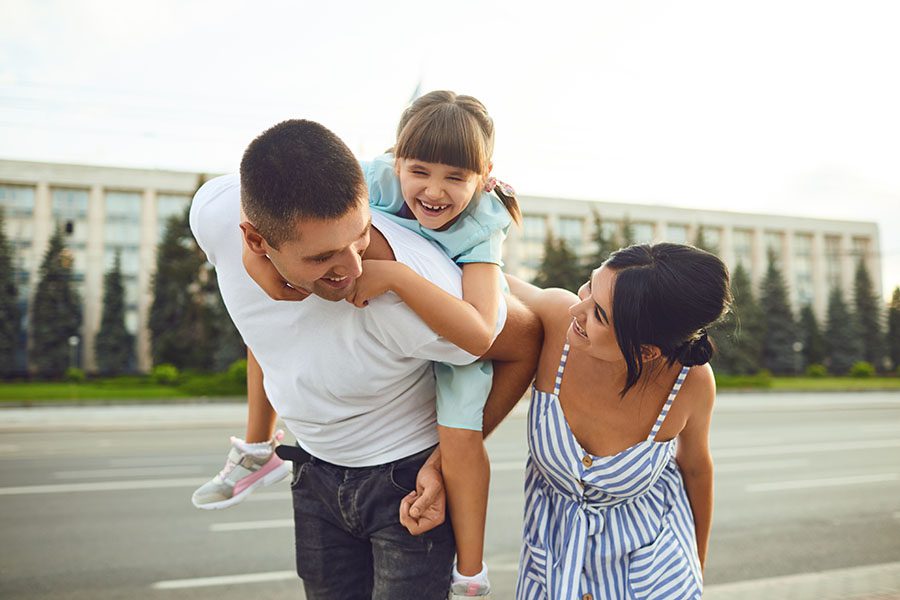 About Our Agency - Cheerful Family Walking Through the City with Dad Giving His Daughter a Piggyback Ride